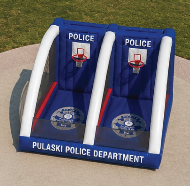 An inflatable police-themed basketball game stand set on a circular concrete court in a grassy park. The inflatable features two blue backboards with white arches, basketball hoops, and 'POLICE' and 'PULASKI POLICE DEPARTMENT' text.