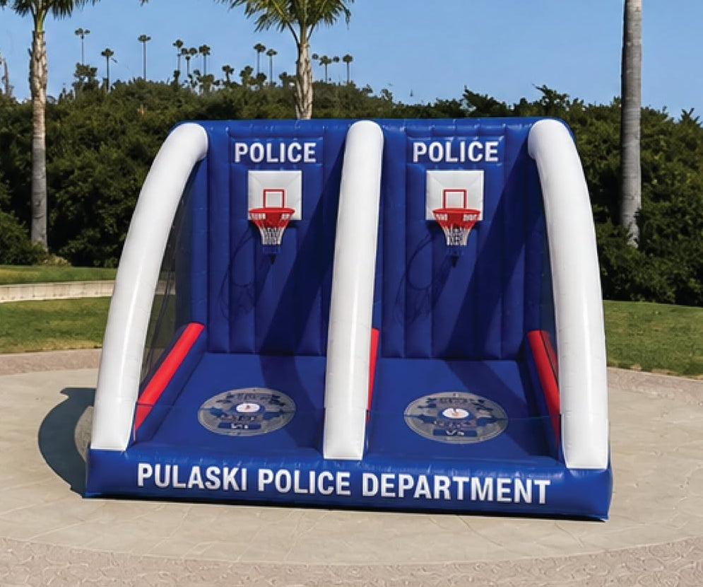 An inflatable double basketball game stand set up outdoors on a grassy lawn. The blue and white inflatable features two basketball hoops with 'POLICE' and 'PULASKI POLICE DEPARTMENT' text on the front.